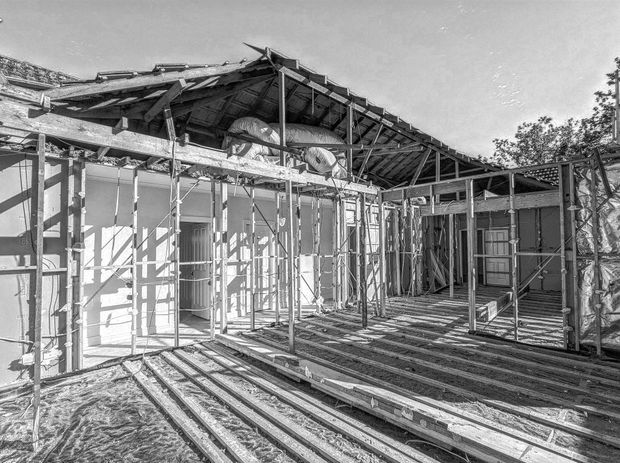 Black-and-white photograph of a single-storey residential building under construction, showing exposed timber framing, roof structure, scaffolding, insulation, unfinished wall linings, and a partially formed deck or floor area.