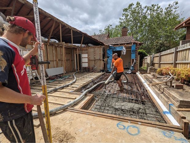 Construction site showing workers preparing reinforcement mesh and formwork for a concrete slab or foundation. Timber framing, plumbing conduits, construction equipment, and an adjacent residential structure are visible outdoors under an overcast sky.