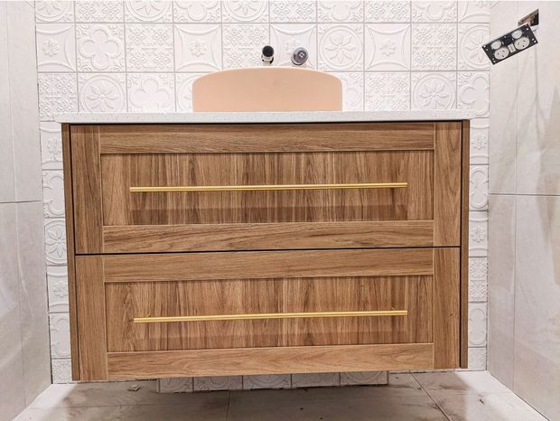 A compact bathroom vanity with two large wood-grain drawers and slim brass-gold handles, topped by a pale benchtop and a peach-toned basin. The vanity is set against decorative embossed white wall tiles, with pale side wall tiles and unfinished-looking floor visible below.