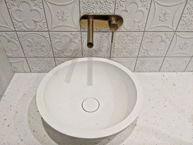 Close-up view of a bathroom vanity featuring a white round vessel basin on a light terrazzo-style benchtop, with a brushed brass wall-mounted tap and an ornate embossed patterned tile splashback.