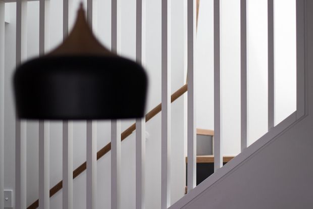 Interior view of a contemporary stairway seen through closely spaced white vertical balusters. A timber handrail rises diagonally behind the balustrade, with a dark pendant light in the foreground and a bright, minimal stairwell beyond.