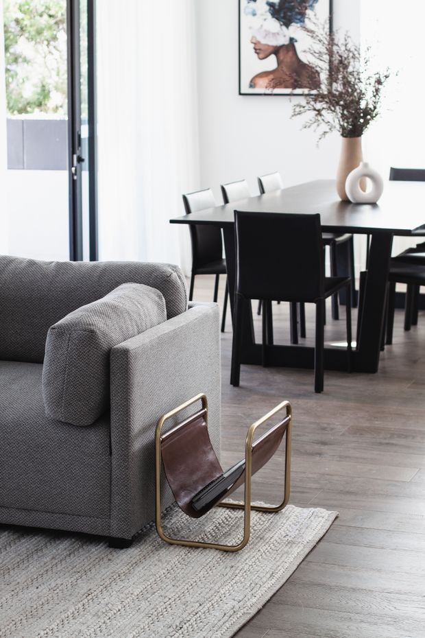 A bright, refined open-plan interior combining a contemporary living area and dining space. A textured grey sofa sits on a light woven rug in the foreground, with a sculptural leather-and-brass lounge chair nearby. Behind it, a black dining table with matching black chairs is positioned beside full-height glazing. White walls, pale wood flooring, minimalist artwork, and a ceramic vase with dried branches complete the neutral scheme.