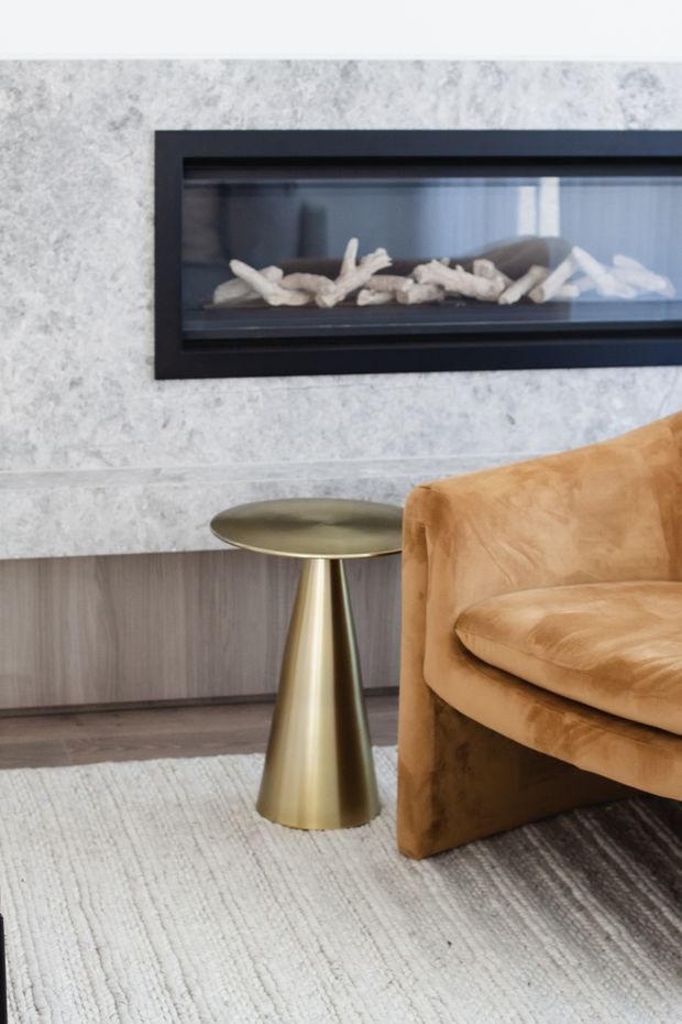 Close-up interior detail of a refined living room featuring a linear black-framed fireplace set into a light textured stone wall, a sculptural brushed-gold pedestal side table, a caramel-brown upholstered armchair, dark timber cabinetry, and a pale textured area rug.