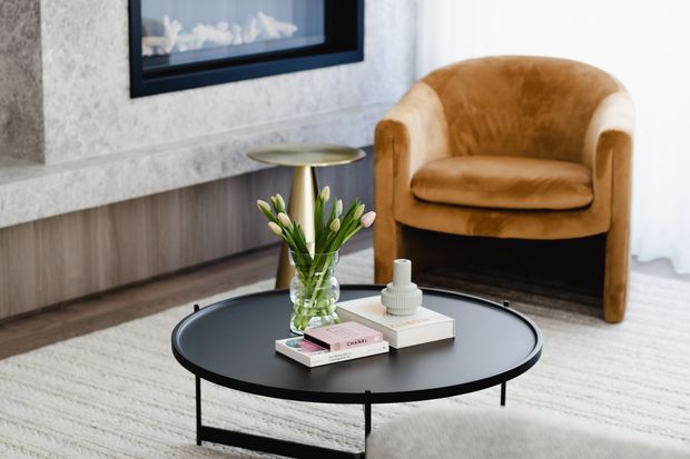 A refined living room vignette featuring a low round black coffee table with books, a vase of flowers and decorative objects, paired with a tan upholstered armchair. A textured light stone fireplace or media wall with timber-toned lower joinery forms the backdrop, with sheer curtains and a pale rug adding softness.