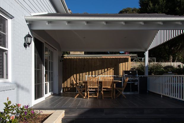 A covered outdoor entertaining area attached to a white-painted brick house, with a dark tiled or paved floor, timber dining table and chairs, built-in barbecue, timber privacy fencing, and white balustrades. The roofed terrace is brightly sunlit and suited to alfresco dining.
