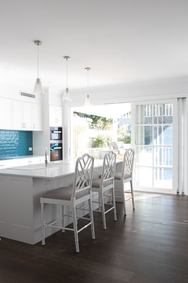 Bright, airy kitchen with a white island and extensive white cabinetry, turquoise glazed-tile splashback, dark timber flooring, and three pendant lights. A row of stools lines the island, with a glazed door and windows opening toward an outdoor area.