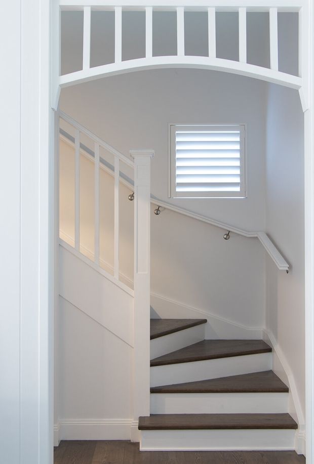 A bright, narrow residential stairway viewed through a doorway, featuring dark timber stair treads with white painted risers, white wall panelling, a decorative curved balustrade overlooking the stairs, a white handrail with metal brackets, and a shuttered window. The space has a refined traditional character with clean white detailing and a neutral palette.