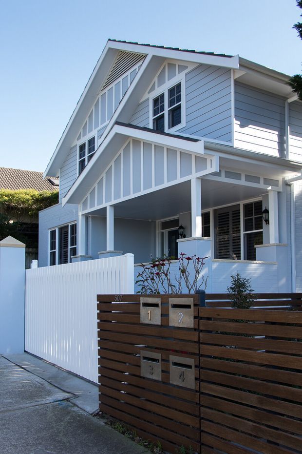 Street-facing view of a narrow multi-storey residential house with a light blue-grey weatherboard facade, prominent gabled roof, decorative white trim, and a covered front porch. A white picket-style boundary fence and dark horizontal timber-slat mailbox gate occupy the foreground.