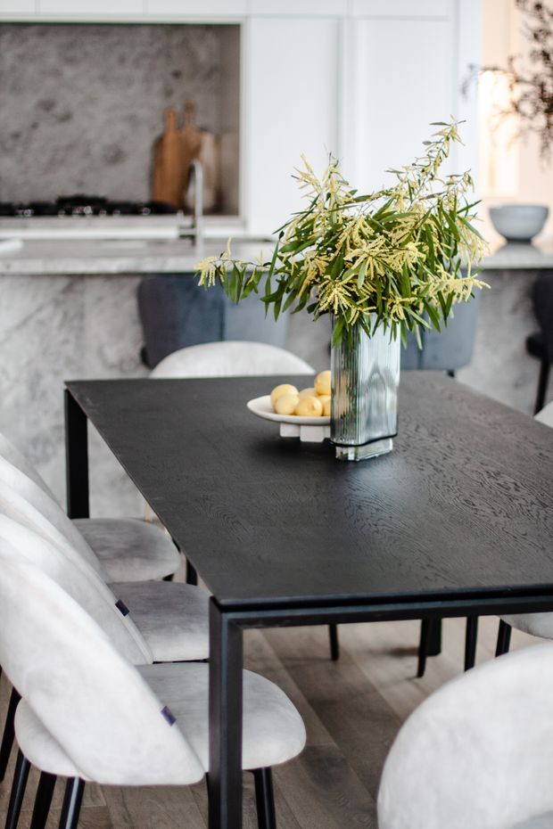 A refined dining area with a dark timber dining table surrounded by upholstered light-grey chairs. A tall ribbed glass vase with leafy branches forms the centerpiece, with a shallow fruit bowl beside it. In the background is a contemporary kitchen featuring pale stone surfaces, white cabinetry, a dark stone splashback, and a slim island or counter.