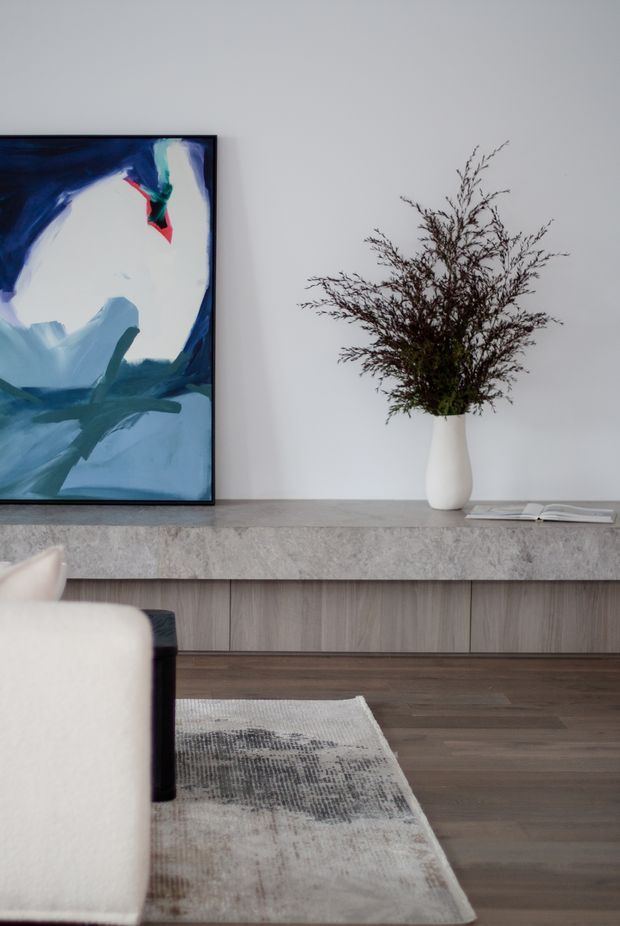A refined contemporary living room vignette featuring a low built-in timber-grain media console with a grey stone benchtop, white painted wall, dark timber flooring, and a neutral textured rug. An abstract blue artwork, sculptural white vase with branches, and the edge of a cream upholstered sofa complete the calm, minimalist arrangement.