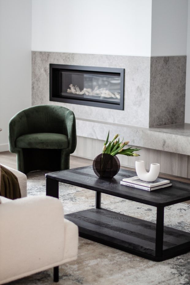 A refined contemporary living room centred on a long black rectangular coffee table with a lower shelf. The room features a linear built-in fireplace set into a pale grey stone or concrete-look surround, a deep green upholstered lounge chair, a light neutral sofa, and a muted patterned rug. A dark round vase with greenery and a sculptural white vessel sit on stacked books, creating a restrained, sophisticated palette.
