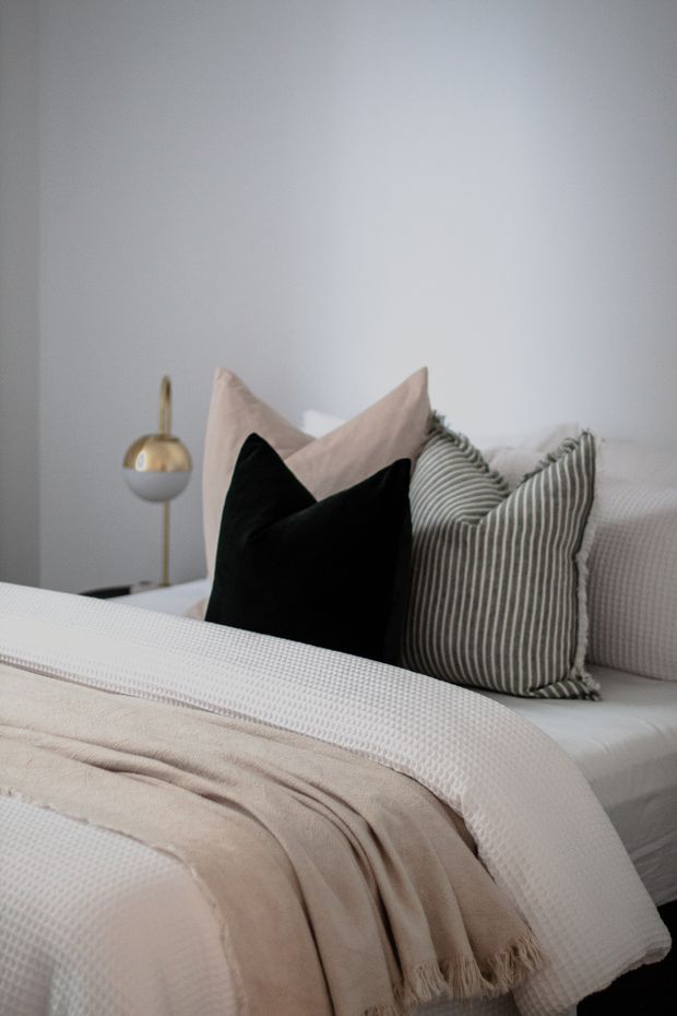 A softly styled bedroom featuring a neatly made bed with layered white and cream textured bedding, a beige throw blanket, and decorative pillows in black, blush beige, and muted grey stripes. A small brass and white bedside lamp sits against a plain light-grey wall, creating a calm, refined neutral interior.