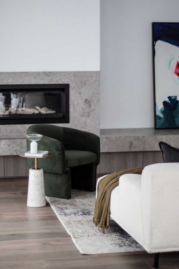 A refined contemporary living room featuring a linear black fireplace set into a pale grey stone-clad wall, dark timber cabinetry, and a low stone hearth shelf. A deep green upholstered armchair sits beside a small round side table, while a white boucle sofa with a draped neutral throw occupies the foreground. A patterned area rug, timber flooring, and abstract artwork complete the calm, modern interior.