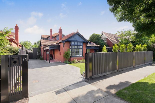 Street-facing view of a red-brick, single-storey home with a prominent gabled entry, dark timber detailing, multiple brick chimneys, and a tiled pitched roof. The property has a broad concrete driveway, landscaped front garden, and a contemporary dark vertical-slat sliding gate and fence.