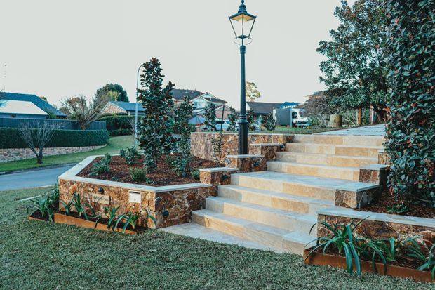 A landscaped residential front entry with broad stone steps, raised garden beds, retaining walls, and a prominent traditional-style lamp post. The hardscape uses warm beige and tan stone with darker natural stone accents, surrounded by manicured lawn and planting. Suburban homes and hedges are visible in the background.