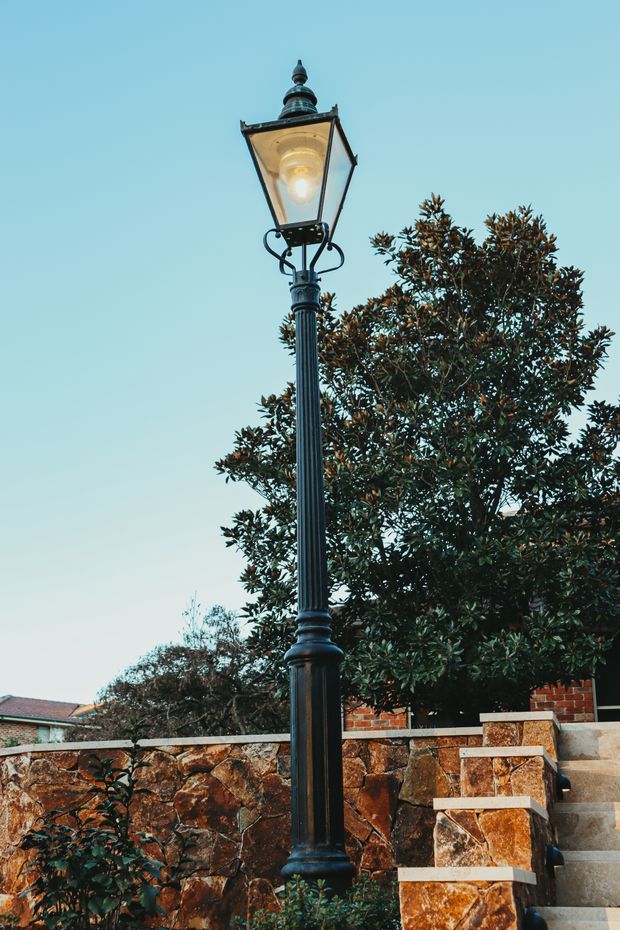 A tall black ornamental street lamp with a traditional lantern-style fixture stands beside a landscaped stone retaining wall and outdoor steps. Mature greenery and part of a residential property are visible in the background under a clear blue sky.
