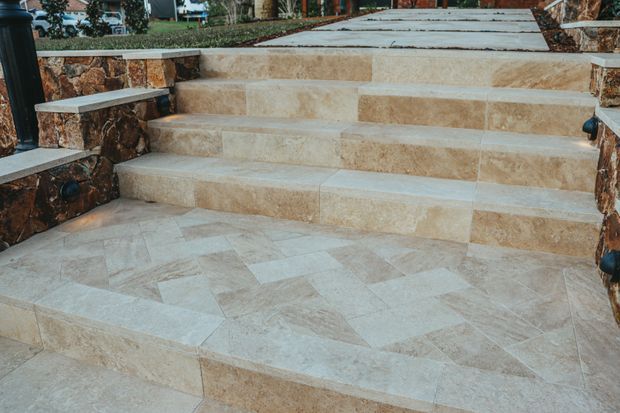 Exterior stepped entrance or terrace finished in light beige natural stone, with broad tiled steps, patterned stone paving, and rustic stacked-stone retaining walls and side benches. The image focuses on hard landscaping and an outdoor circulation area.
