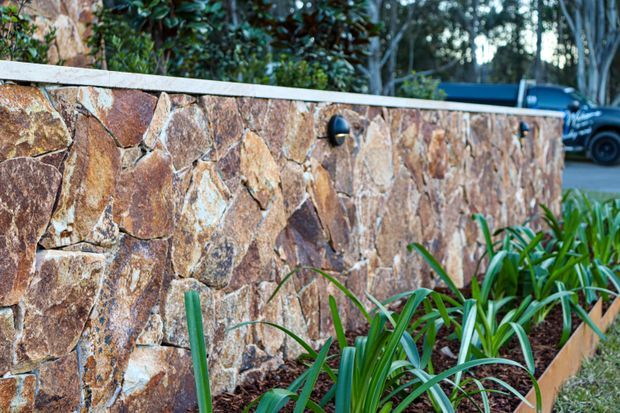 Close-up exterior view of a rough natural stone retaining or boundary wall with a pale stone cap, integrated dark wall light, and landscaped planting bed in front. Dense greenery and part of a driveway with a parked vehicle are visible in the background.