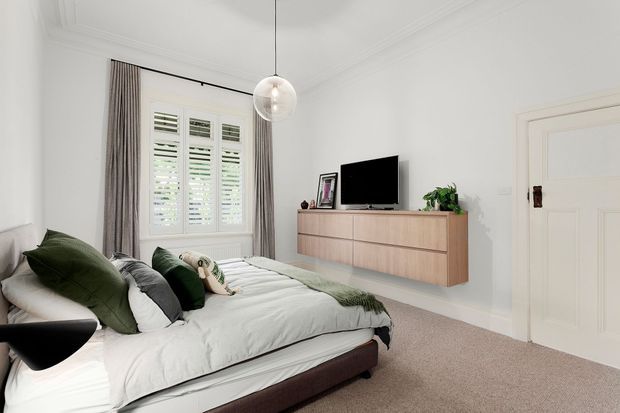 Bright, refined bedroom with a low upholstered bed, layered white and green bedding, floating timber-toned dresser, wall-mounted television, indoor plant, pendant globe light, plantation shutters, and full-height curtains. The room has white walls with decorative cornice detailing and a soft neutral carpet.