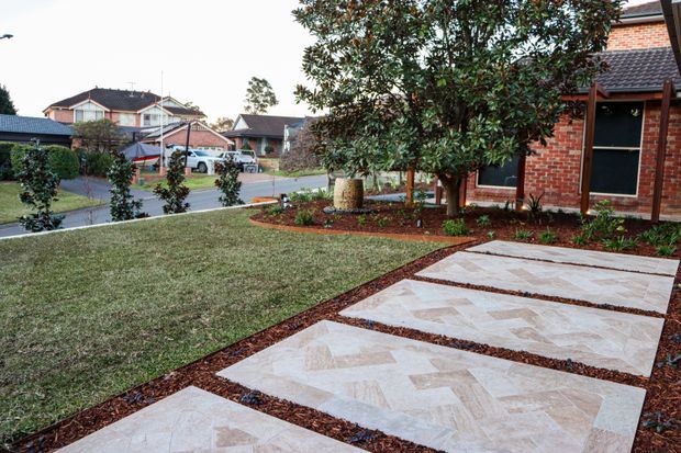 A landscaped suburban front yard beside a red-brick home, featuring a broad lawn, planted garden beds, mature trees, and a geometric paved walkway made from pale stone slabs with dark mulch joints. Neighbouring residential homes and a street are visible beyond the garden.