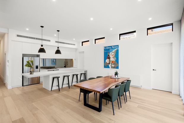 Bright open-plan kitchen and dining area with white cabinetry, a long island with bar stools, pale timber flooring, and a timber dining table with upholstered chairs. High-level windows, recessed ceiling lights, and black pendant lights create a clean contemporary interior. A colourful framed poster provides a small visual accent.