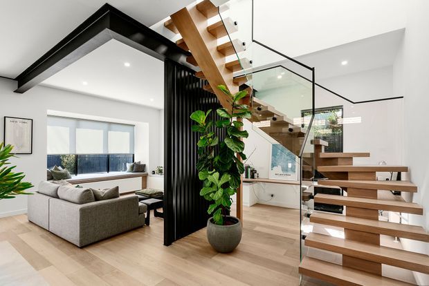Bright contemporary open-plan interior featuring a timber floating staircase with clear glass balustrades, black metal framing and a vertical black slatted screen. The staircase connects the living area to an upper level, with pale timber flooring, white walls, recessed lighting, indoor planting and a neutral sectional sofa. A large skylight or clerestory opening adds natural light above the stair.