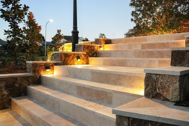 An outdoor landscaped terrace or garden feature with broad stepped stone seating or stairs, integrated warm lighting, and low stone retaining walls. Planting and a decorative black lamp post frame the scene at dusk.