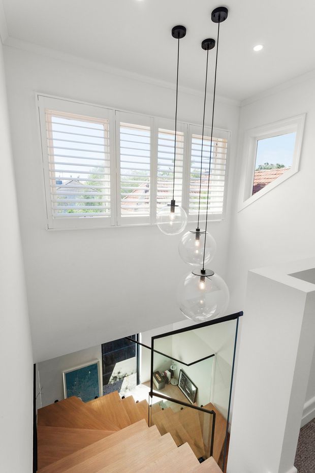 Bright contemporary stairwell with a timber staircase, clear glass balustrade, black metal handrail, and a cluster of spherical pendant lights. White walls and ceiling surround shuttered windows, while a triangular window provides views of neighbouring rooftops. The stair opening offers a partial view to the lower level.