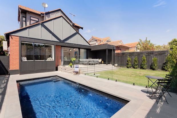 A renovated suburban home with a prominent gabled extension clad in dark grey panels and red brick, opening onto a timber entertaining deck and rectangular swimming pool. The rear garden includes lawn, fencing, shrubs, and a small outdoor dining setting. Terracotta tiled roofs of neighbouring homes are visible beyond the property.