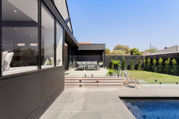 Contemporary residential rear courtyard viewed from a paved pool terrace, featuring dark cladding, extensive floor-to-ceiling glazing, a raised timber-toned deck with outdoor seating, a lawn, fencing, and a swimming pool. The architecture has strong rectilinear lines and a deep roof overhang.