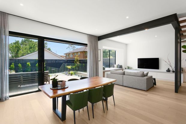 A bright open-plan dining and living area with pale timber flooring, a long timber dining table, green upholstered dining chairs, and a light grey sectional sofa facing a wall-mounted television. Full-height glazing opens to a balcony or terrace, while dark structural framing and sheer curtains add contrast. The space has a clean contemporary fit-out and overlooks neighbouring suburban homes and greenery.