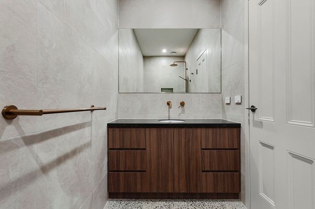 Compact contemporary bathroom featuring full-height light grey stone-look wall tiles, a dark stone benchtop with wall-mounted brass tapware, and a wall-hung dark timber vanity with integrated basin. A large frameless mirror reflects the shower area, while white decorative-panel doors and a timber towel rail add contrast. The floor has a small-format grey and white patterned tile finish.