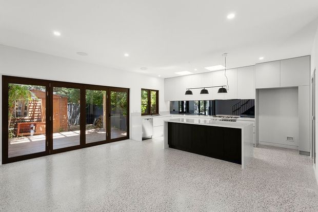 Bright open-plan kitchen and living area with a large white island, dark timber cabinetry, integrated appliances, black pendant lights, white walls, and polished speckled concrete or terrazzo-style flooring. Dark-framed glazed doors open to an outdoor deck, while a stairway is visible at the rear.