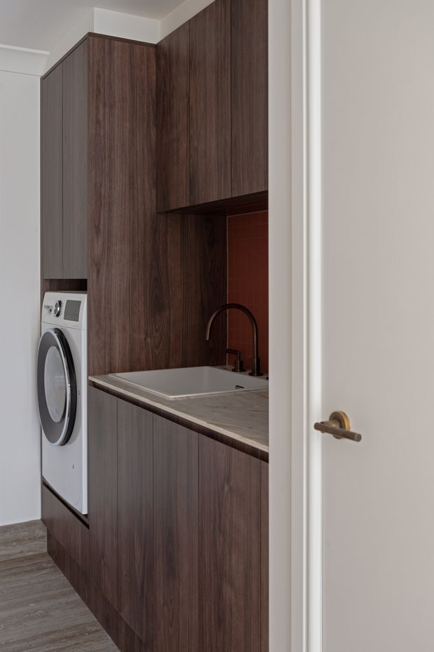 Compact laundry room viewed through an open white door, with dark wood-grain built-in cabinetry, an integrated front-loading washing machine, a rectangular utility sink, dark curved faucet, grey stone-look benchtop, and a small reddish-brown tiled splashback. The room has a narrow, highly functional built-in layout and grey wood-look flooring.