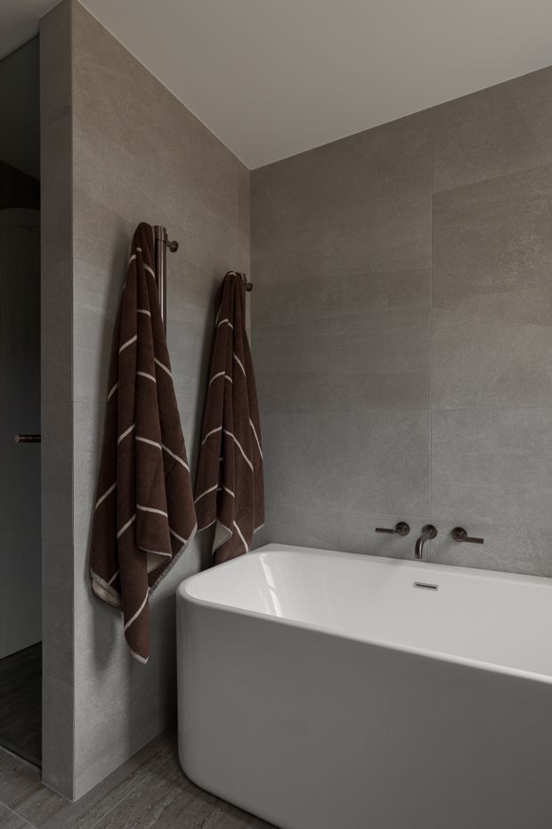 A contemporary bathroom featuring a freestanding white bathtub, large-format grey stone-look wall tiles, grey tiled flooring, and dark wall-mounted tapware. Brown striped towels hang on the tiled partition wall, and a partial shower area is visible at left.