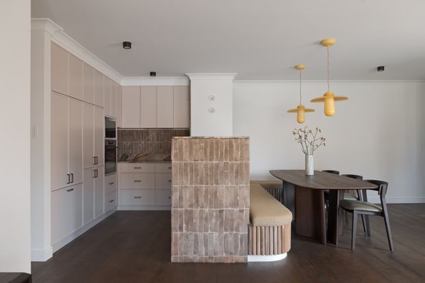 A refined open-plan kitchen and dining area with full-height pale cabinetry, integrated appliances, a tiled kitchen island with a curved upholstered breakfast bench, and a dark oval dining table with matching chairs. The space features dark timber flooring, white walls and ceiling, classic cornice detailing, and sculptural mustard-gold pendant lights.