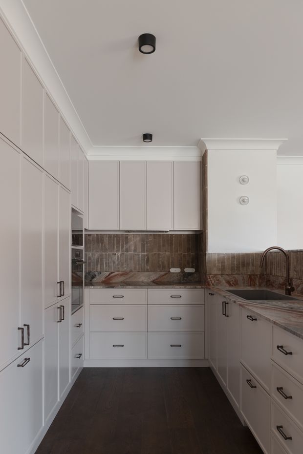 A refined galley kitchen with extensive off-white shaker-style cabinetry, dark timber flooring, and a dramatic veined stone benchtop and splashback. The kitchen includes tall pantry-style storage along the left wall, integrated appliances, a sink beneath a raised decorative bulkhead, dark metal pulls, and black cylindrical downlights. A restrained neutral palette is complemented by the warm brown and reddish veining of the stone surfaces.