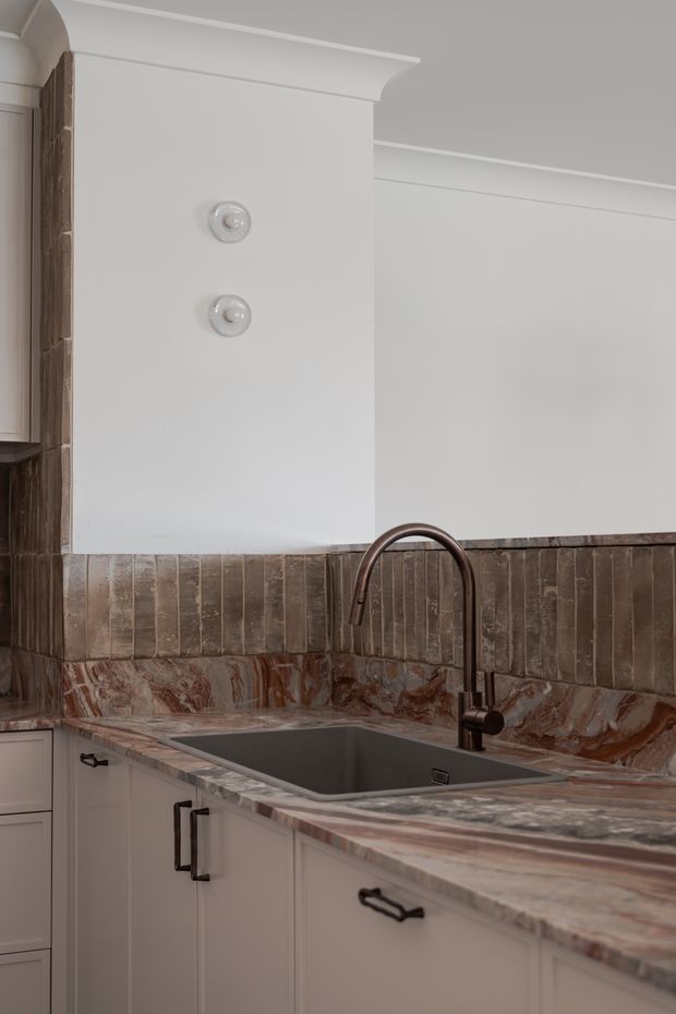 Close-up view of a refined kitchen corner featuring pale painted cabinetry with dark metal handles, a deep undermount sink and curved dark faucet, and a dramatic veined stone benchtop. The backsplash is clad in vertically arranged dark brown textured tiles, with matching stone continuing along the wall. White walls feature decorative cornice detailing and two circular wall fixtures.