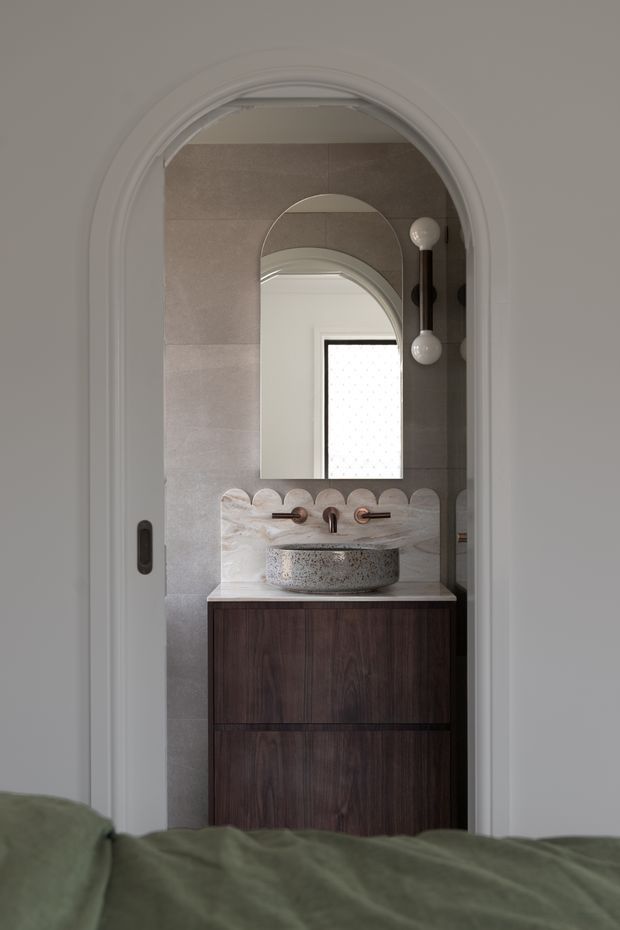 A compact bathroom vanity viewed through an arched doorway from a bedroom. The bathroom features large taupe-grey wall tiles, a dark timber floating vanity, a pale stone benchtop with an integrated or vessel-style stone basin, copper-toned wall-mounted tapware, and an arched mirror. Decorative scalloped stone detailing runs along the vanity splashback, with a sculptural wall light beside the mirror. The surrounding doorway has ornate white trim, while the foreground shows part of a muted green bed.