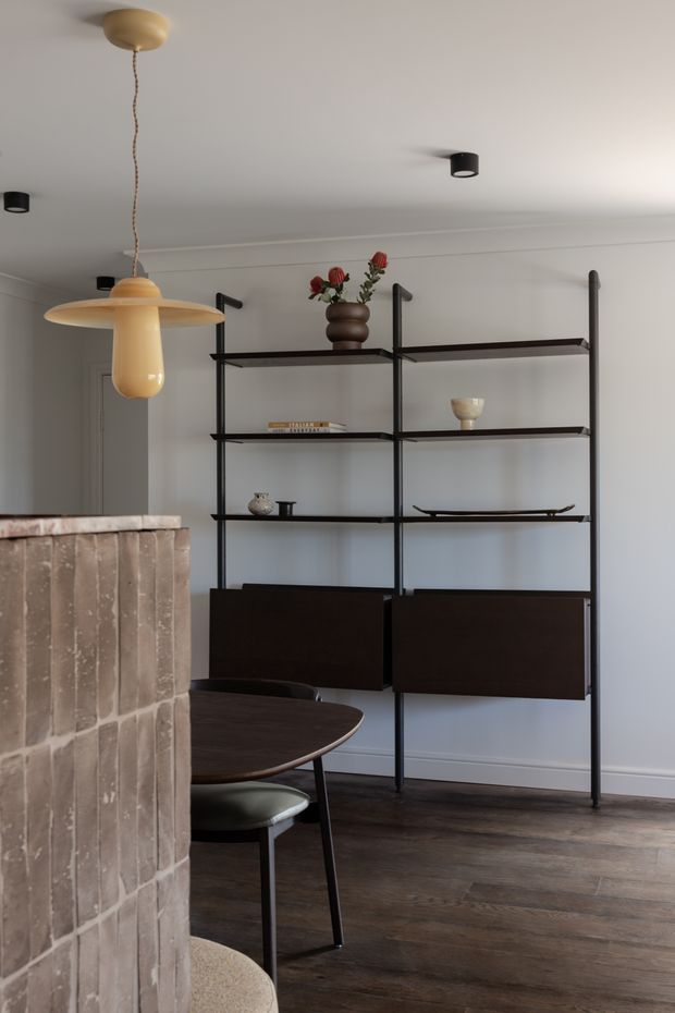 A refined dining area viewed from behind a pale textured brick fireplace or partition. The room features a dark timber dining table and chairs, a freestanding black metal shelving and storage unit, warm sculptural pendant lighting, dark timber flooring, and white walls and ceiling. Minimal decorative objects and muted furnishings create a contemporary, calm atmosphere.