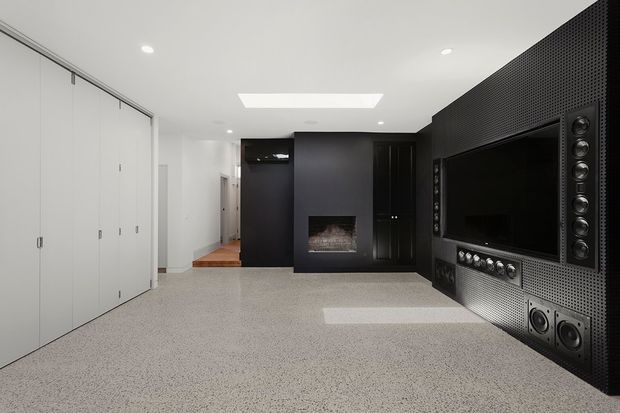 A sleek, dark home media room with a large built-in television and surround-sound speaker wall, a central fireplace, extensive white storage cabinetry, recessed ceiling lighting, and a light speckled floor. The space has a minimalist, high-contrast contemporary design.