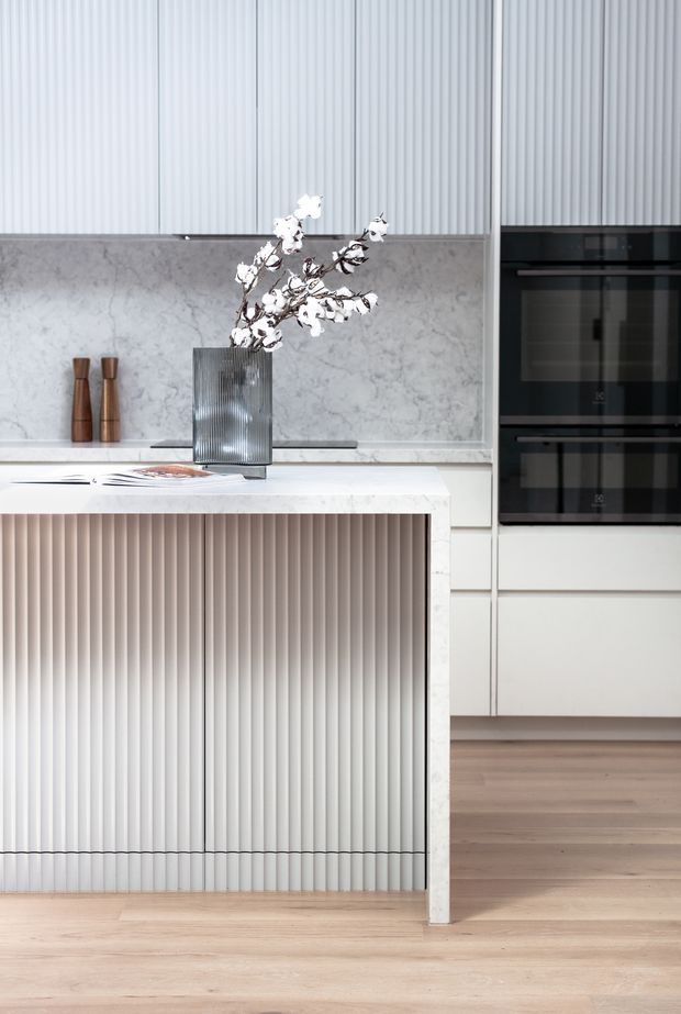 A bright contemporary kitchen featuring a large island with vertically fluted white cabinetry and a pale stone benchtop. The rear wall has a matching stone splashback, tall pale cabinetry, ribbed wall detailing, and integrated black double ovens. Light timber flooring and restrained styling create a refined, minimal look.