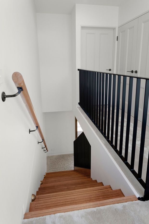 A bright, narrow residential stairway viewed from the upper landing, with warm timber treads, white walls and trim, a black vertical metal balustrade, and a timber handrail mounted on the wall. White paneled doors and carpeted upper and lower landings are visible.