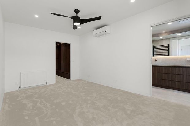 Bright, unfurnished bedroom with soft beige patterned carpet, white walls and ceiling, recessed downlights, and a black three-blade ceiling fan. A dark timber doorway opens to a hallway or wardrobe area, while a glazed opening reveals an adjoining bathroom with dark timber vanity cabinetry, pale stone benchtop, wall-mounted towel radiator, and mirrored cabinetry.