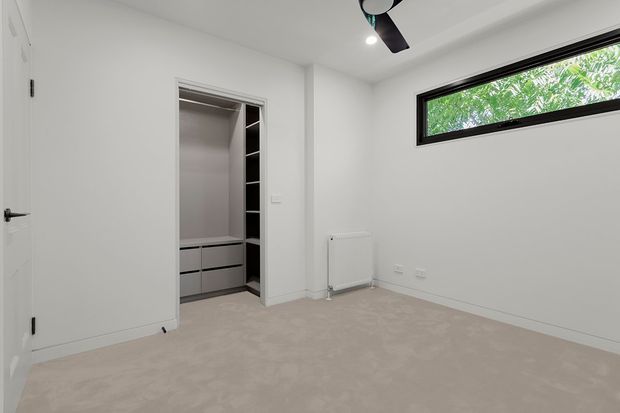 Empty, bright bedroom with white walls, a beige carpeted floor, a built-in open wardrobe, a narrow horizontal black-framed window, a white radiator, and a ceiling fan. The room has a clean, contemporary minimalist appearance.