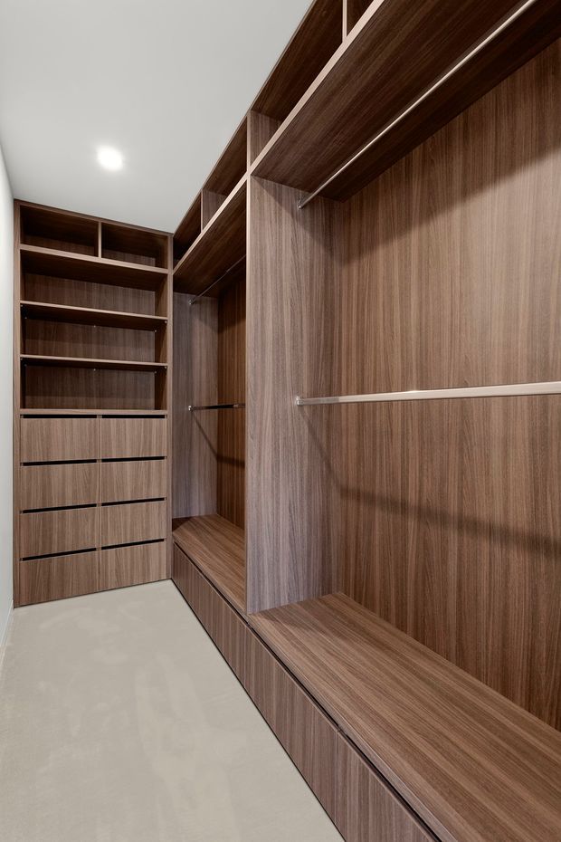 A custom walk-in wardrobe with extensive dark wood-grain built-in cabinetry, open shelving, drawers, hanging rails, and a long integrated bench or storage ledge. The room has a clean white ceiling with recessed lighting and a light neutral floor, creating a streamlined contemporary fitted-storage interior.