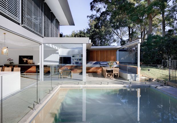 A bright contemporary residential pool courtyard viewed from the pool edge. The scene features a glazed indoor-outdoor living area, white rendered walls and slim structural frames, horizontal external blinds, timber-lined outdoor seating and dining zones, a covered terrace, and a glass pool fence. Mature trees and a fenced garden sit beyond the entertaining area.