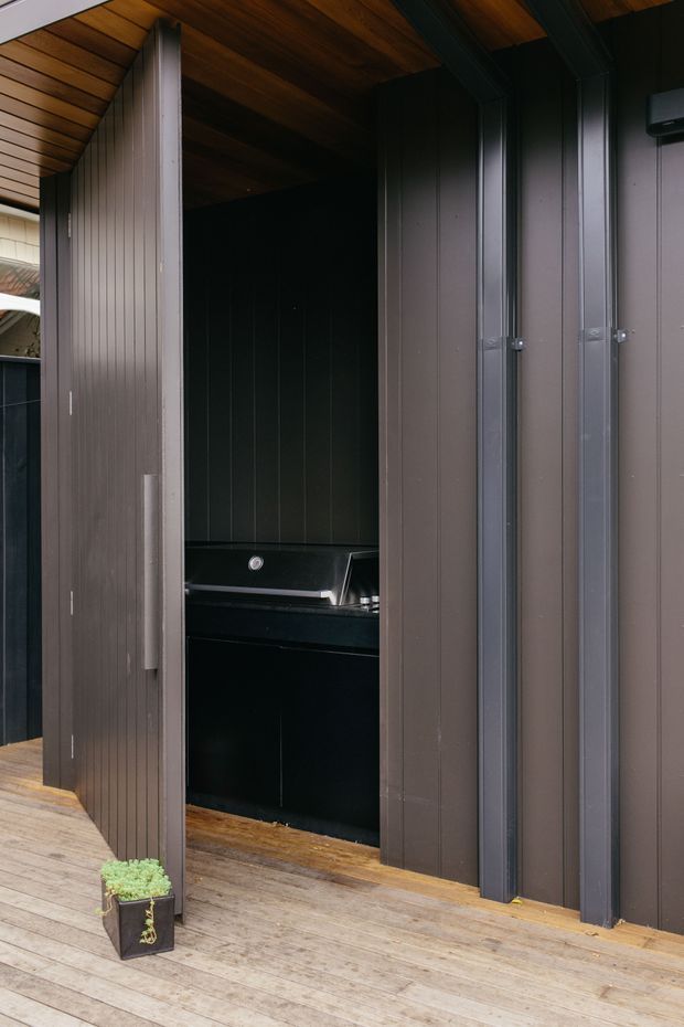 A compact outdoor cooking area set into a dark vertical-panelled enclosure, with a built-in black barbecue and countertop beneath a warm timber-lined ceiling. The space opens onto a timber deck, with sliding or folding vertical-panel doors framing the cooking zone.