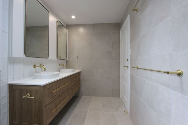A bright, contemporary bathroom with a long double-sink timber vanity, white countertop, rounded-rectangle mirrors, pale large-format wall and floor tiles, and brushed brass tapware, drawer pulls, towel rail, and shower hardware. A white door and recessed ceiling light complete the clean, refined space.
