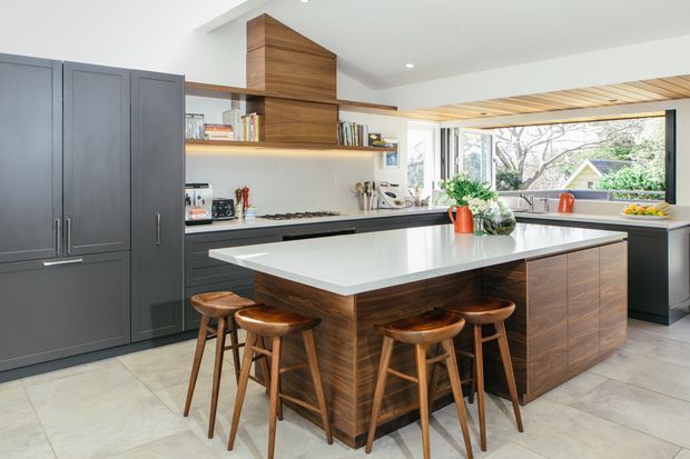 A bright, contemporary kitchen with a large white island featuring seating for four, dark grey full-height cabinetry, warm timber island joinery, open shelving, and a timber-lined ceiling detail. Large windows provide views to greenery and neighbouring homes.